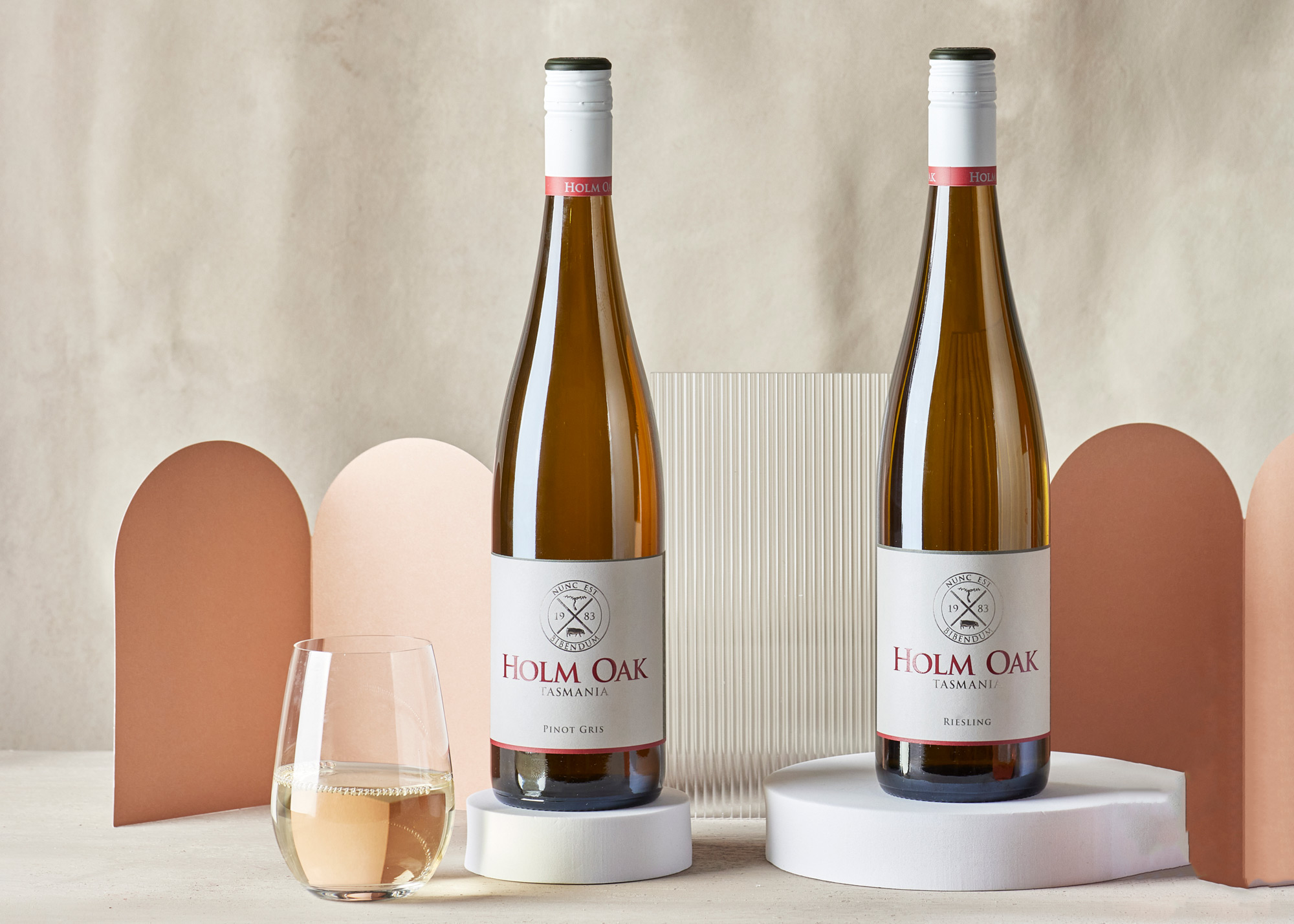 A bottle of Holm Oak Riesling and a bottle of Holm Oak Pinot Gris on plinths with a stemless glass of white wine on a table